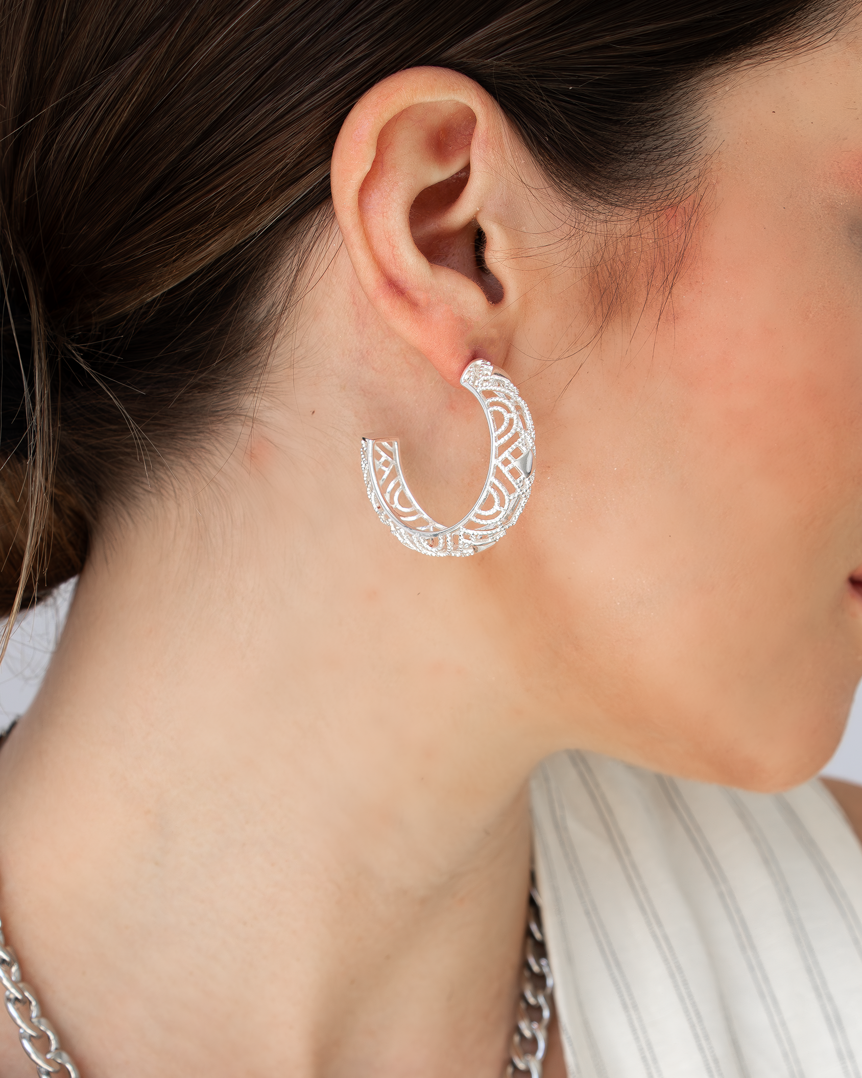 A close-up image of a woman's ear wearing a silver hoop earring with intricate carvings.
