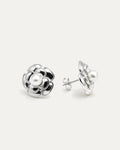 A close-up studio shot of a pair of silver-tone flower stud earrings on a plain white or light beige background. Each earring