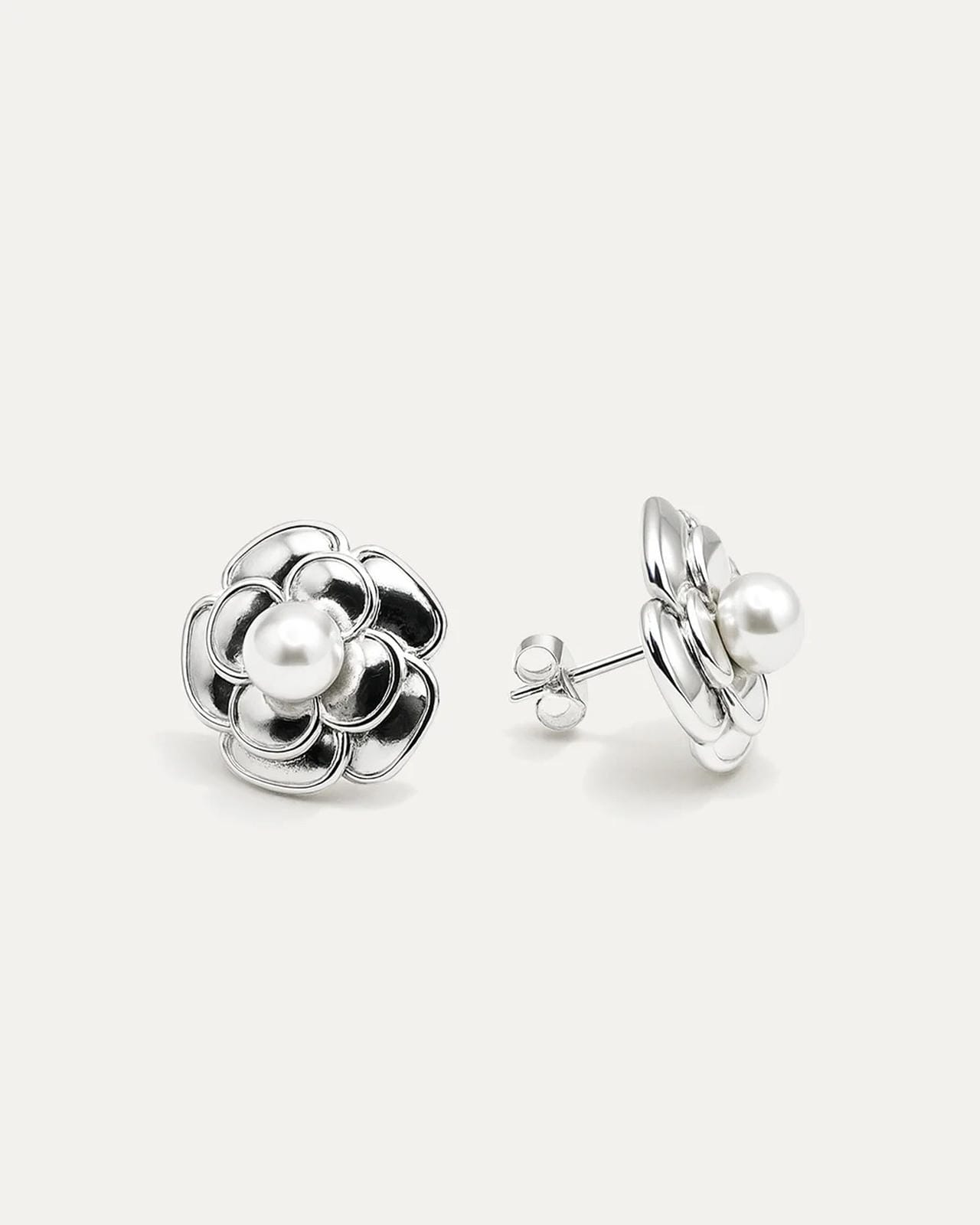 A close-up studio shot of a pair of silver-tone flower stud earrings on a plain white or light beige background. Each earring