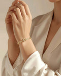 A model wearing a white silk blouse, showcasing a gold-plated chain bracelet on her wrist. The bracelet features rectangular