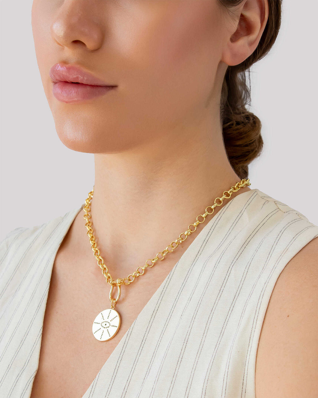 Gold pendant necklace with engraved sunburst coin charm, worn with a light blouse.
