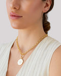 Gold pendant necklace with engraved sunburst coin charm, worn with a light blouse.