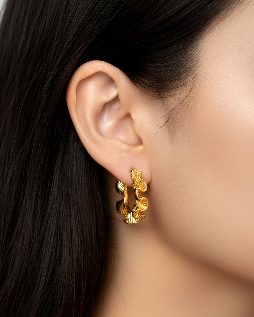 A close-up studio shot of a pair of small, thick gold-tone hoop earrings on a plain white or light beige background.
