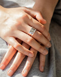 Close-up of a woman's hands resting on grey fabric, highlighting an intricate silver-tone 'Aurora Band Ring' with a central r