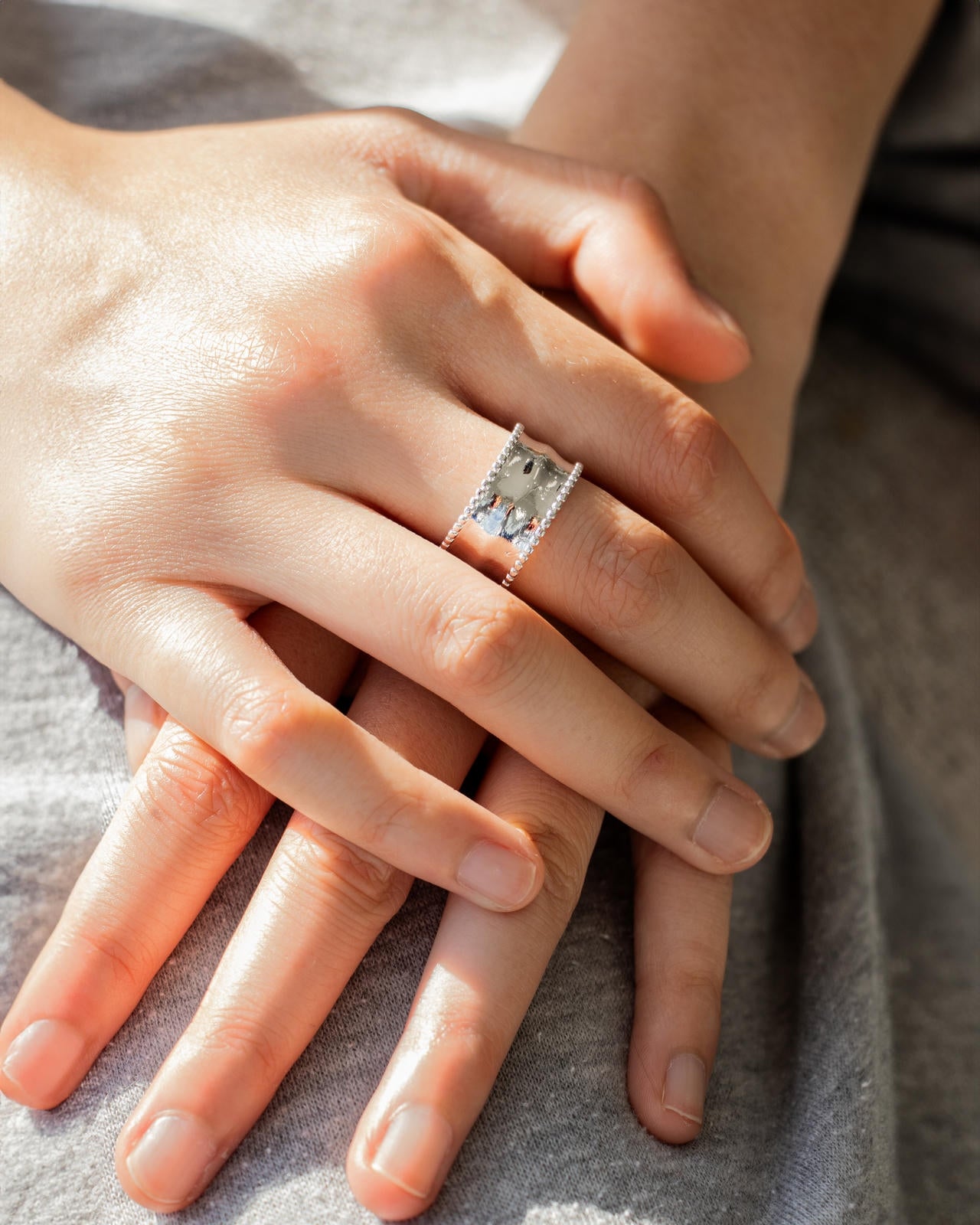 Close-up of a woman's hands resting on grey fabric, highlighting an intricate silver-tone 'Aurora Band Ring' with a central r