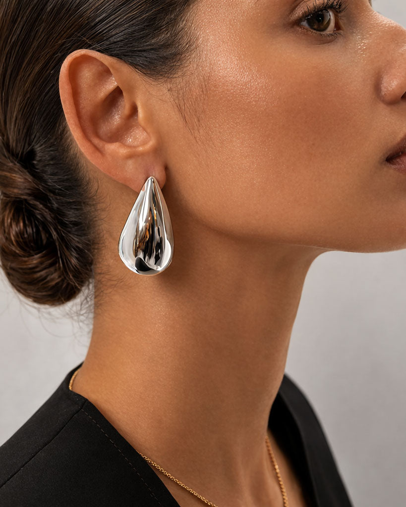 A pair of silver bold teardrop stud earrings with smooth sculptural surface, displayed against a soft neutral background.