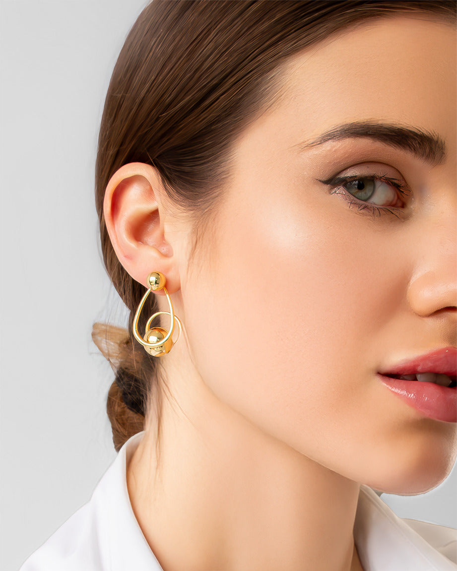 Gold sculptural drop earrings with overlapping loops and sphere accents, worn on model.