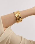 A model's arm wearing a wide, polished gold cuff bracelet. The statement open-cuff features a bold, sculptural design of laye