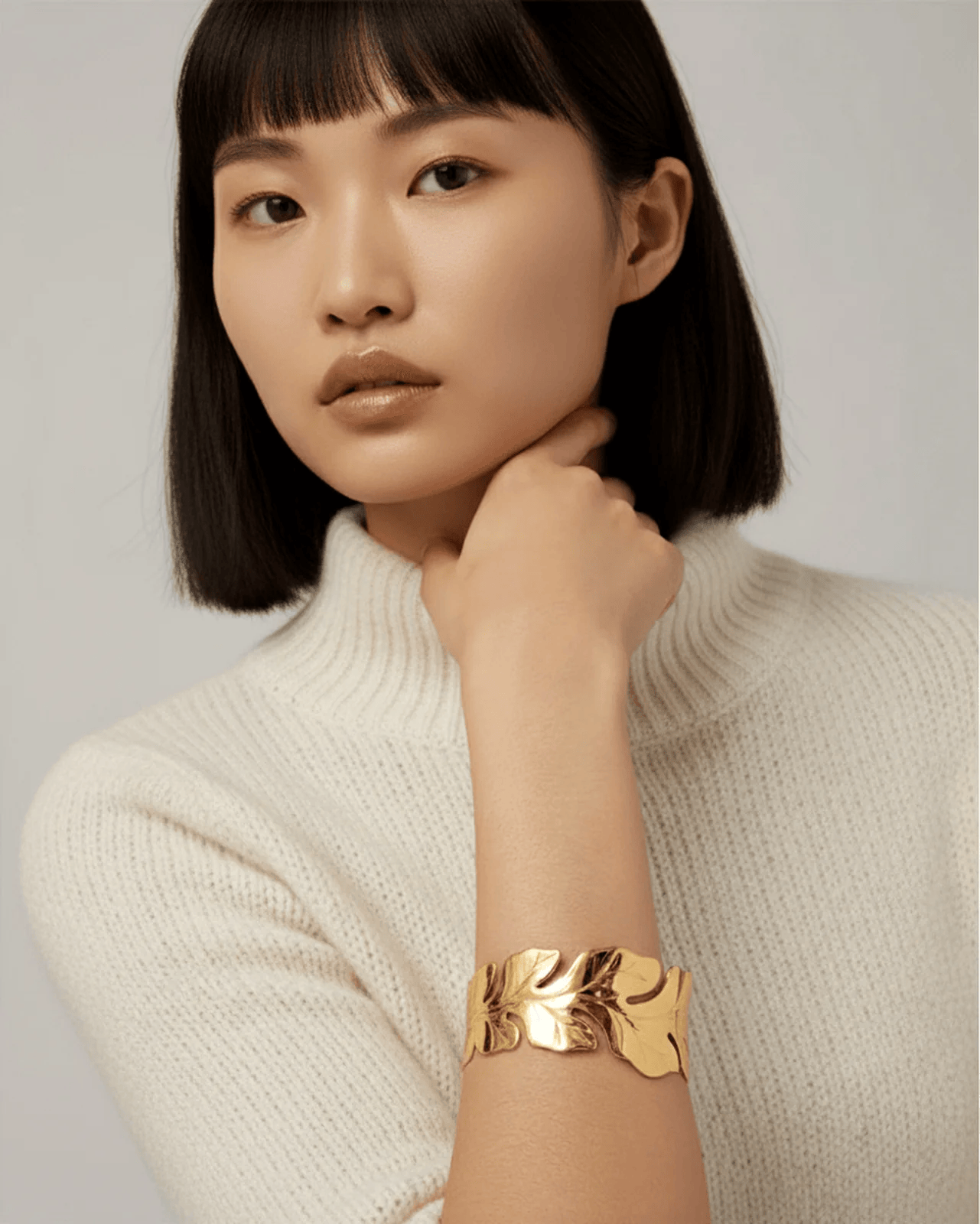 A head-and-torso shot of a model with a dark bob and bangs, wearing a white turtleneck sweater. She is showcasing a wide, pol