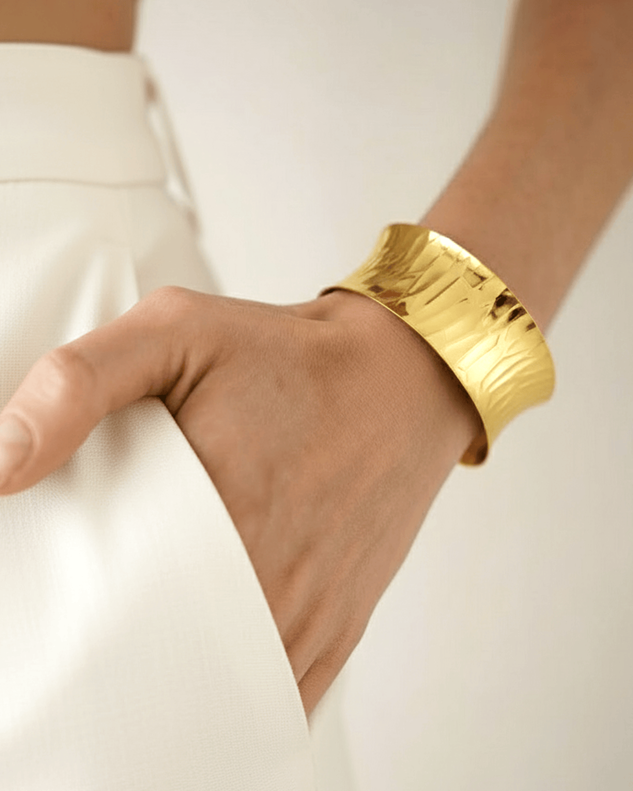 A close-up of a woman's hand in a white pant pocket, showcasing a wide, gold-toned cuff bracelet. The chunky, open-cuff featu