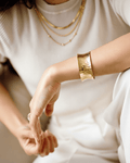 A torso-up view of a woman wearing an all-white outfit and a statement gold cuff bracelet. The wide, sculptural open-cuff has