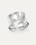 A wide, polished silver open-cuff bracelet with a sculptural, concave shape. The statement cuff features a reflective hammere
