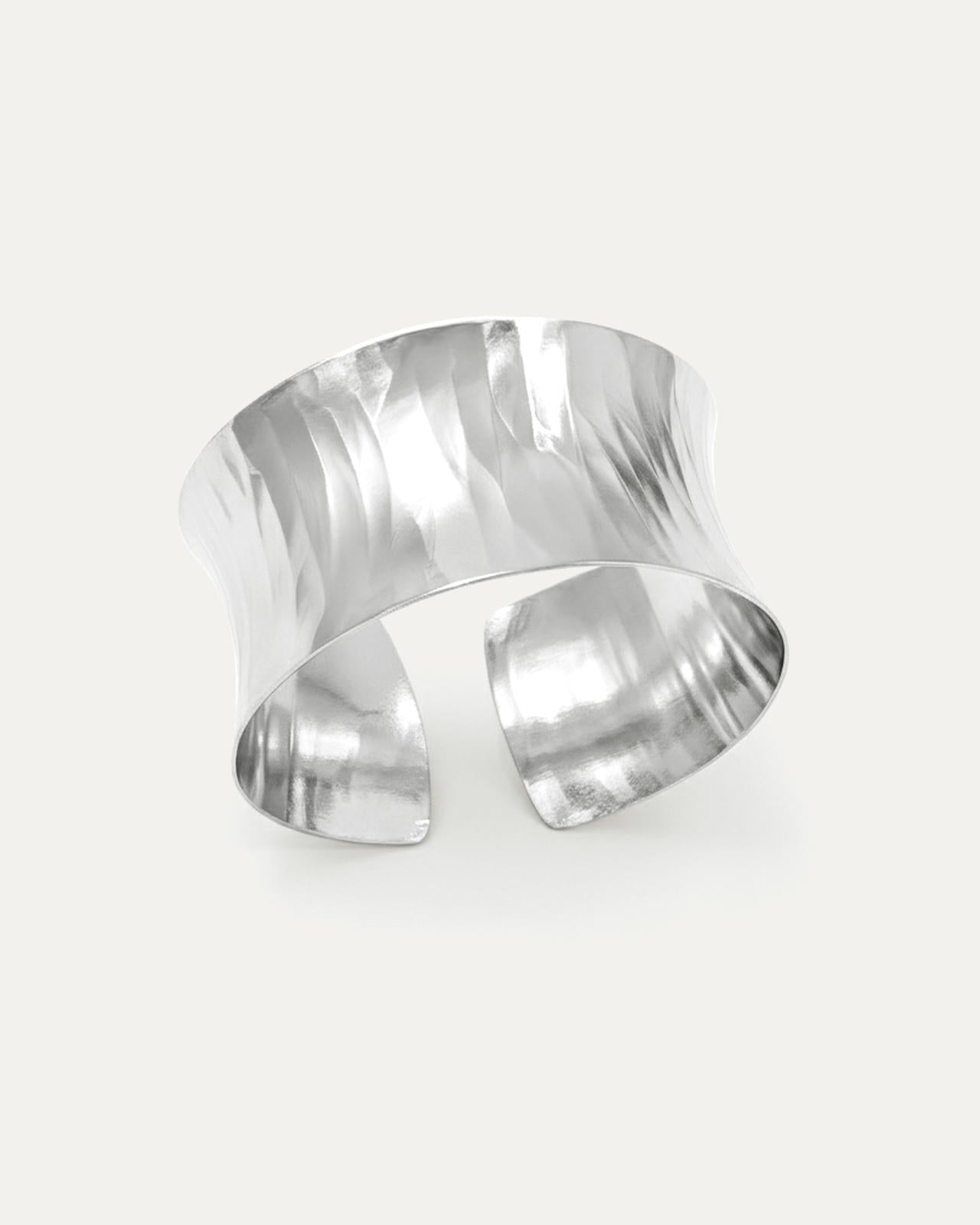 A wide, polished silver open-cuff bracelet with a sculptural, concave shape. The statement cuff features a reflective hammere