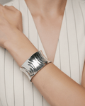 A model wearing a white pinstripe outfit, showcasing a wide, concave silver cuff bracelet. The sculptural open-cuff features 