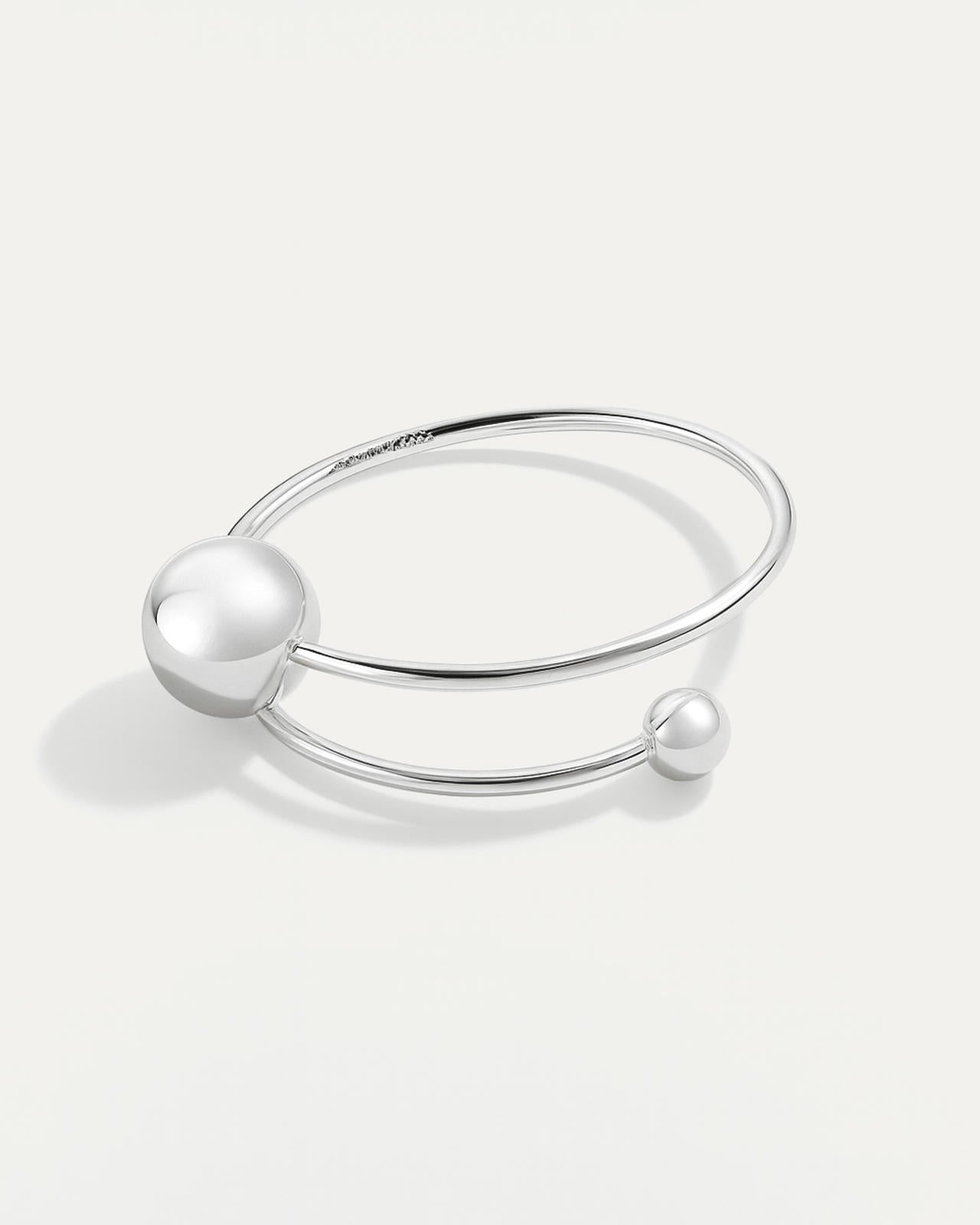 A minimalist, open silver cuff bracelet with a smooth band. The band wraps around, terminating in two polished spheres of con