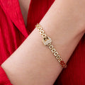 Gold chain-link bracelet with crystal-studded lock clasp shown on wrist against red textured fabric, styled with delicate necklace