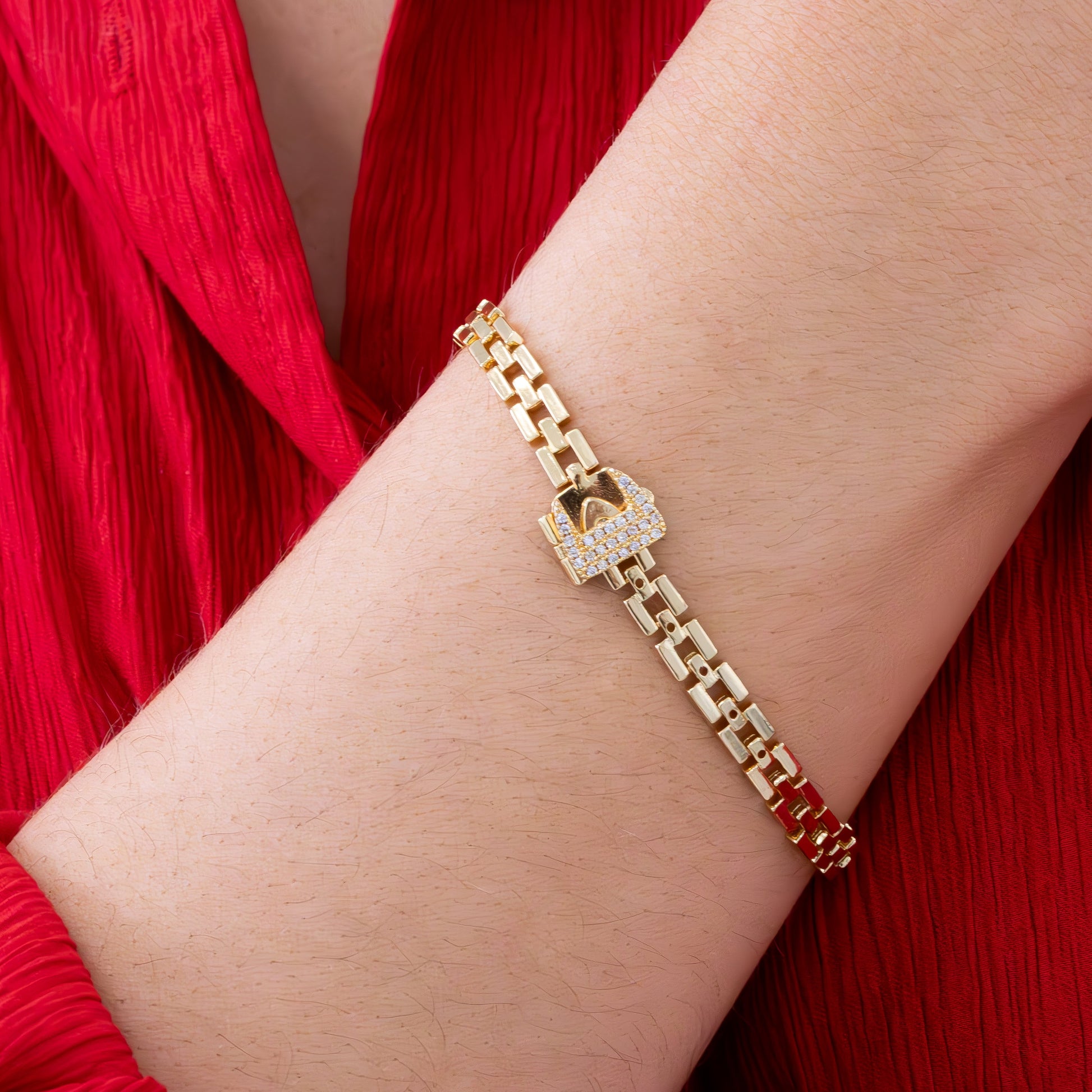 Gold chain-link bracelet with crystal-studded lock clasp shown on wrist against red textured fabric, styled with delicate necklace