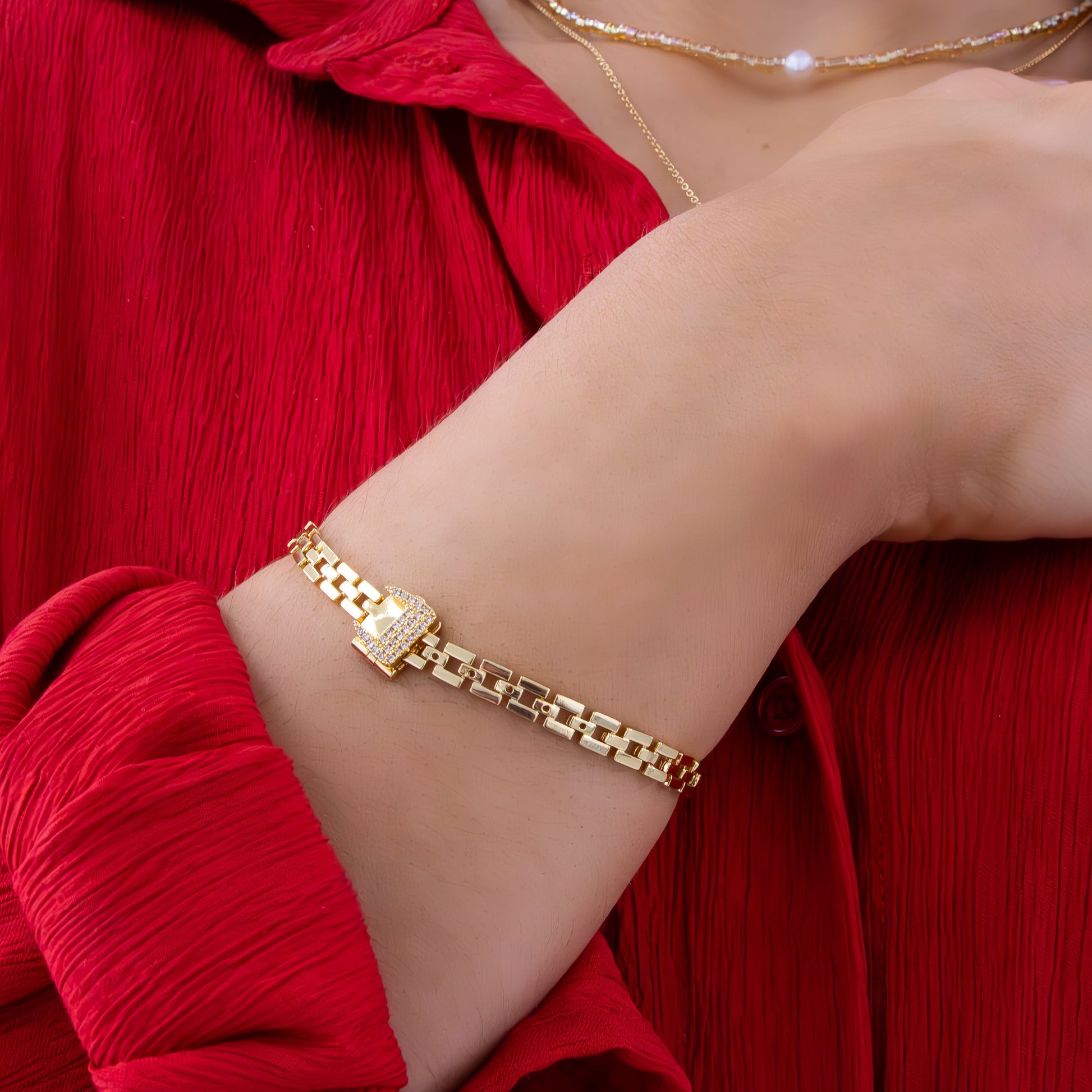 Gold chain-link bracelet with crystal-studded lock clasp shown on wrist against red textured fabric, styled with delicate necklace