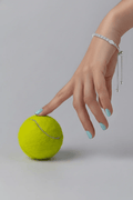 A model's hand with light blue nail polish reaching down to touch a neon yellow tennis ball. The wrist wears a silver-plated 