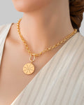 Gold pendant necklace with engraved sunburst coin charm, worn with a light blouse.