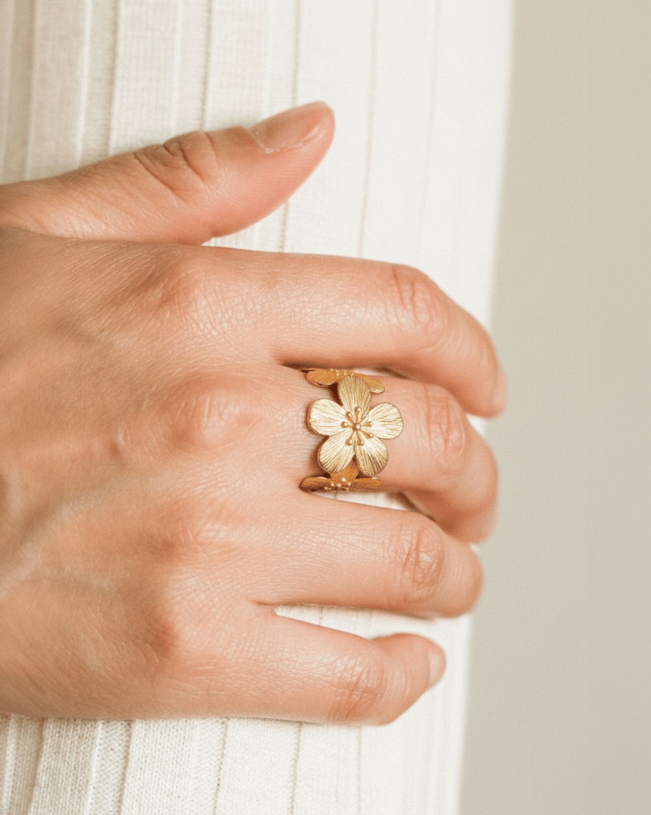 A close-up of a hand wearing a wide gold-tone floral ring on the index finger. The ring features a large, textured flower des