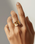A close-up photograph features a person's hand wearing a bold, sculptural gold-tone ring on their index finger.  
