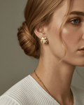 A close-up portrait of a woman features a gold-tone flower stud earring on her earlobe. The earring has layered, textured pet