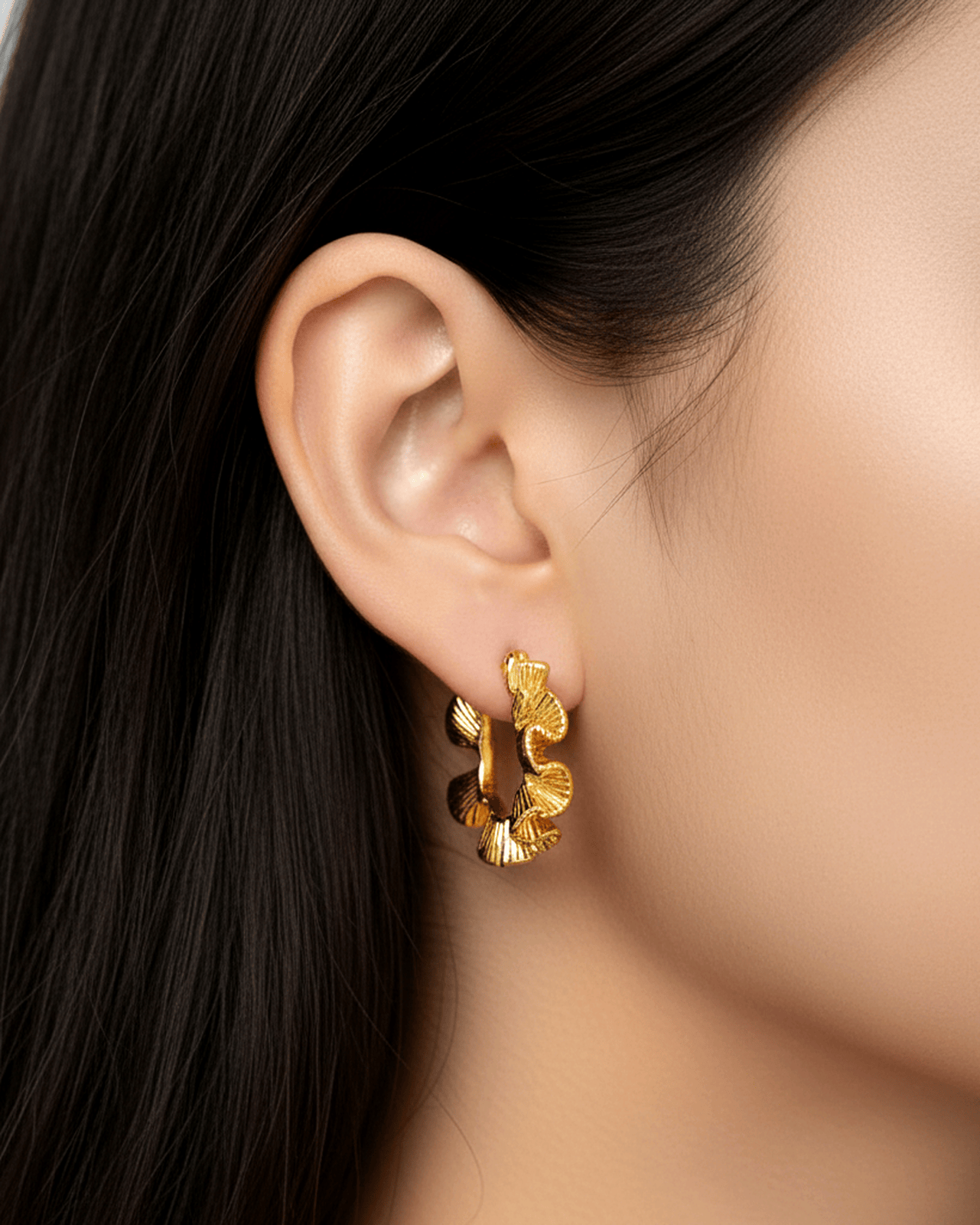 A close-up photograph of a woman's ear shows a small, thick gold-tone hoop earring worn on the lobe.  