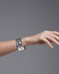 A model's wrist wearing a wide, polished silver open-cuff bracelet. The statement cuff features a pronounced uneven, hammered